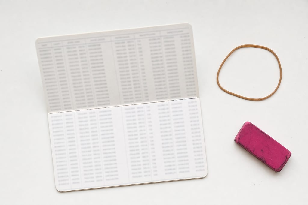 A small savings passbook lying open on a white desk showing ruled entry columns, a loose rubber band and a worn pink eraser next to it